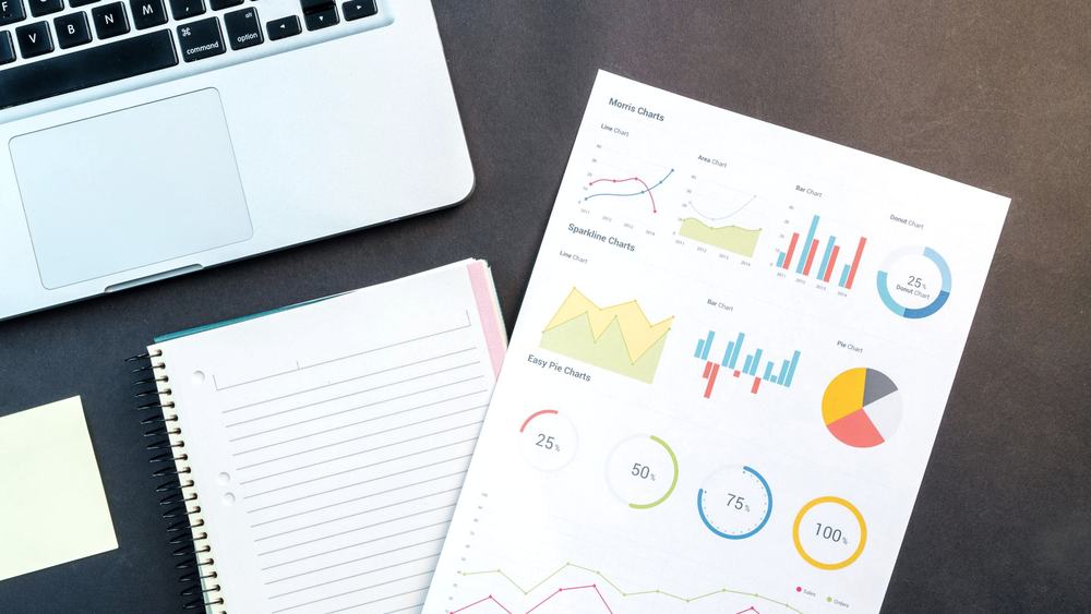 A printed financial report with charts and summary figures lying beside a laptop and notebook.