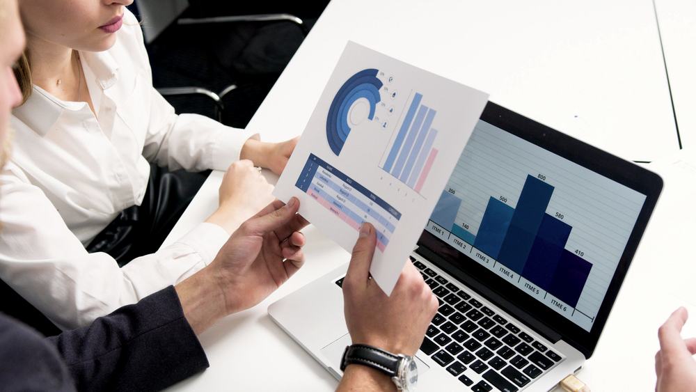 A person reviewing a printed performance chart beside a laptop showing financial figures.