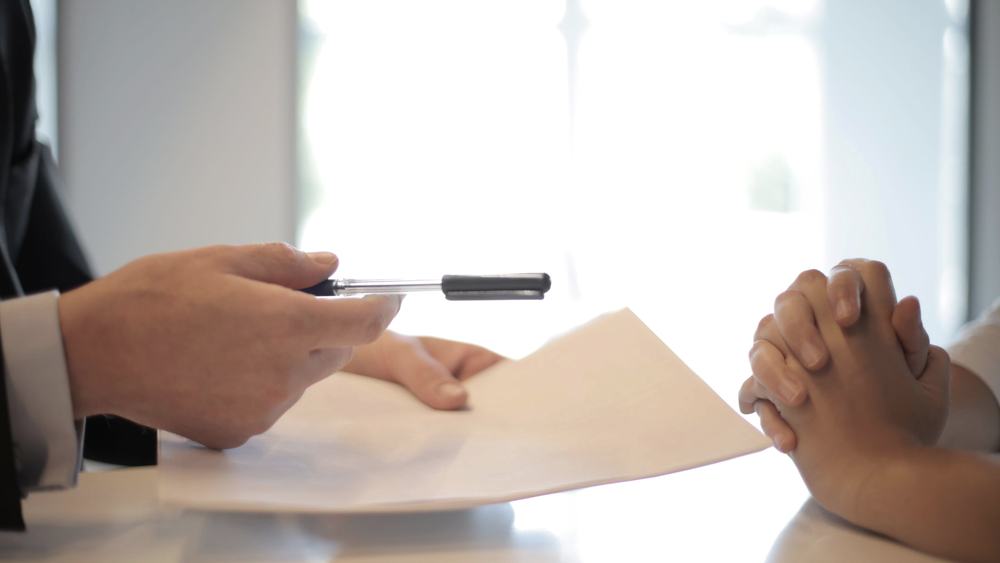 Two people seated across a desk, one holding a pen over a document ready to sign.